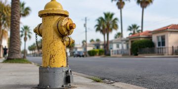 Low-angle close-up of a fire hydrant with a locked steel cover over its bolts on a street of stucco houses and palm trees in Southeast Los Angeles.