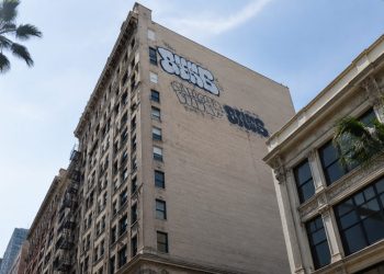Low-angle view of a tall historic masonry building in downtown Los Angeles with large spray-painted tags high on its side wall in neutral daylight.
