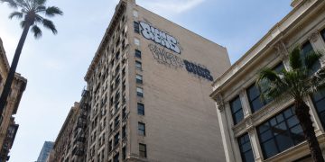 Low-angle view of a tall historic masonry building in downtown Los Angeles with large spray-painted tags high on its side wall in neutral daylight.