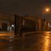 Low-angle view of an industrial street of warehouses in downtown Los Angeles at night, with a steel vehicle gate standing open under street lamps.