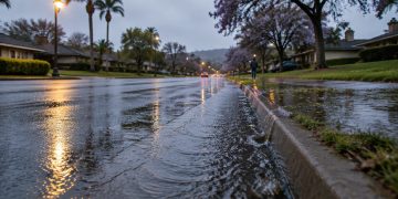 Low-angle view of water running along a wet residential street in Woodland Hills at early-morning twilight, with street lamps lit and houses behind.