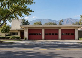 Mid-century single-story fire station with wide bay doors on a Pasadena residential street, with trees and the San Gabriel Mountains behind in daylight.