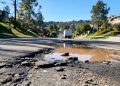 Muddy, wet street in Porter Ranch with broken asphalt and debris in the foreground and a utility truck blurred in the background in afternoon light.