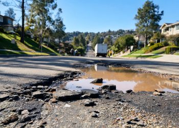 Muddy, wet street in Porter Ranch with broken asphalt and debris in the foreground and a utility truck blurred in the background in afternoon light.