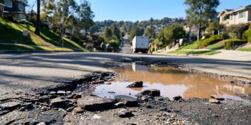Muddy, wet street in Porter Ranch with broken asphalt and debris in the foreground and a utility truck blurred in the background in afternoon light.