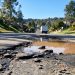 Muddy, wet street in Porter Ranch with broken asphalt and debris in the foreground and a utility truck blurred in the background in afternoon light.