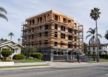 New apartment building under construction beside older single-family homes and palm trees on a Los Angeles street in neutral daylight.