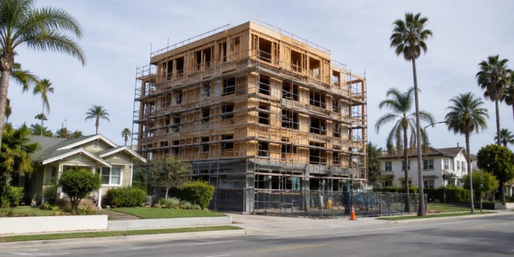 New apartment building under construction beside older single-family homes and palm trees on a Los Angeles street in neutral daylight.