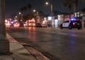 Night street scene on a wide South Los Angeles avenue, with a curb and lamp post in sharp focus and a patrol SUV blurred in the background.