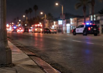 Night street scene on a wide South Los Angeles avenue, with a curb and lamp post in sharp focus and a patrol SUV blurred in the background.