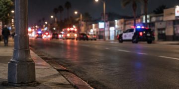 Night street scene on a wide South Los Angeles avenue, with a curb and lamp post in sharp focus and a patrol SUV blurred in the background.