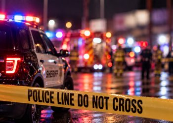 Nighttime emergency response with sheriff and fire vehicles, firefighters and blurred activity behind police tape, with no identifiable storefront visible.