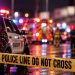 Nighttime emergency response with sheriff and fire vehicles, firefighters and blurred activity behind police tape, with no identifiable storefront visible.