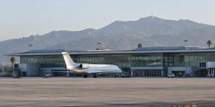 Passenger jet parked on the apron outside a modern airport terminal with a long glass facade in Burbank in soft early-morning daylight.