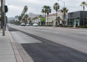 Patched asphalt and a concrete bus pad along Sunset Boulevard in West Hollywood, with palm trees and low-rise buildings in neutral daylight.