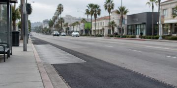 Patched asphalt and a concrete bus pad along Sunset Boulevard in West Hollywood, with palm trees and low-rise buildings in neutral daylight.