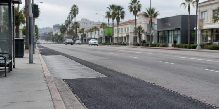 Patched asphalt and a concrete bus pad along Sunset Boulevard in West Hollywood, with palm trees and low-rise buildings in neutral daylight.