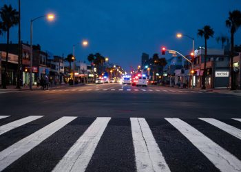 Pre-dawn Long Beach intersection with a marked crosswalk lit by street lamps in the foreground and emergency vehicles blurred far in the background.