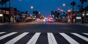 Pre-dawn Long Beach intersection with a marked crosswalk lit by street lamps in the foreground and emergency vehicles blurred far in the background.