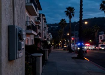 Pre-dawn residential street in Highland Park with a utility meter box in the foreground and emergency vehicles blurred in the distance.
