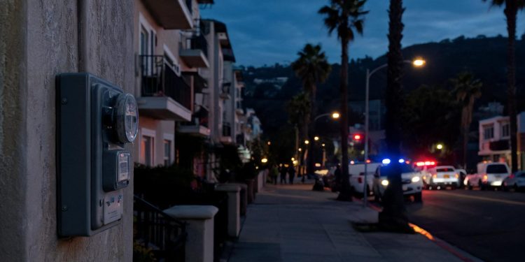 Pre-dawn residential street in Highland Park with a utility meter box in the foreground and emergency vehicles blurred in the distance.