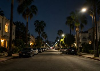 Quiet residential street of apartment buildings and palm trees in West Hollywood at night under street lamps, seen from the middle of the block.