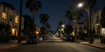 Quiet residential street of apartment buildings and palm trees in West Hollywood at night under street lamps, seen from the middle of the block.