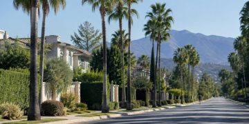 Quiet residential street of large hillside homes and palm trees in Tarzana in neutral morning daylight, seen from a long-lens view down the block.