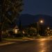 Quiet residential street of older bungalows and mature trees in Monrovia at night under street lamps, with the dark San Gabriel foothills beyond.