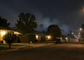 Quiet residential street of single-story homes and mature trees in West Covina at night, with a faint haze of smoke over the rooftops.