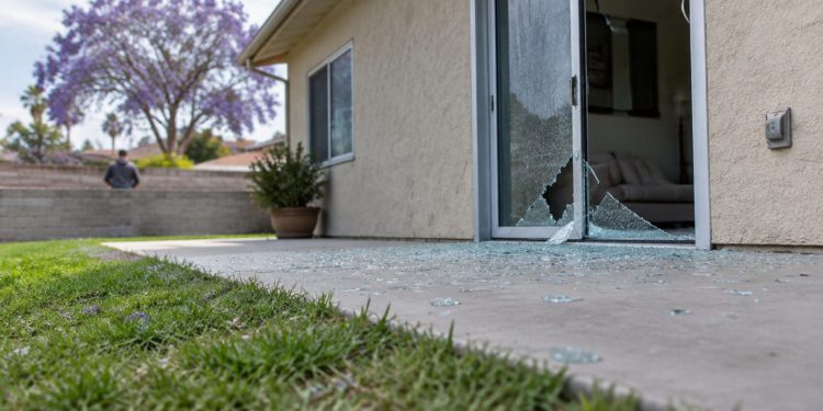 Rear patio of a single-story stucco home in Woodland Hills with a shattered sliding glass door and glass fragments on the concrete in daylight.