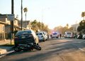 Residential street in Inglewood on a clear afternoon, with a small damaged motorbike beside a parked car and emergency vehicles blurred far behind.