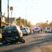 Residential street in Inglewood on a clear afternoon, with a small damaged motorbike beside a parked car and emergency vehicles blurred far behind.