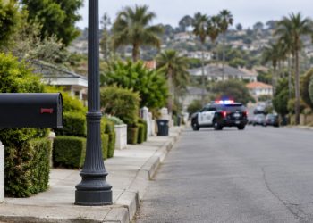 Residential street in Ladera Heights in daylight, with a mailbox and lamp post in the foreground and a patrol SUV blurred in the background.