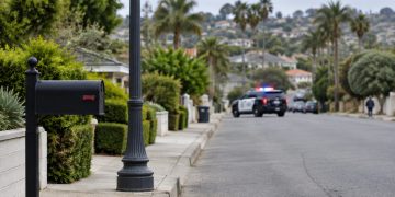Residential street in Ladera Heights in daylight, with a mailbox and lamp post in the foreground and a patrol SUV blurred in the background.