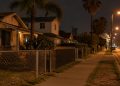 Residential street in Westmont at night with stucco homes, palm trees and an empty sidewalk under street lamps, seen from a long-lens view.