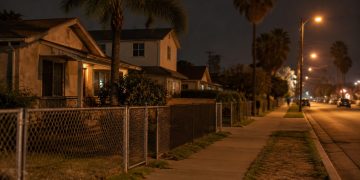 Residential street in Westmont at night with stucco homes, palm trees and an empty sidewalk under street lamps, seen from a long-lens view.