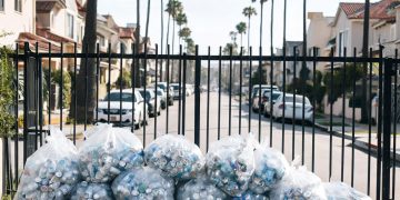 Residential street in the Fairfax District of Los Angeles with trash bags and plastic bottles piled beside a fence, palm trees blurred behind.