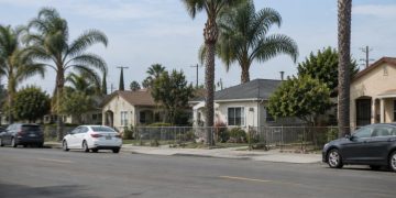 Residential street of single-story stucco homes, front yards and palm trees in Santa Ana, photographed from across the street in neutral daylight.