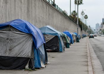 Row of tents along a sidewalk beside a concrete freeway retaining wall in San Diego in neutral daylight, seen with a long lens down the block.