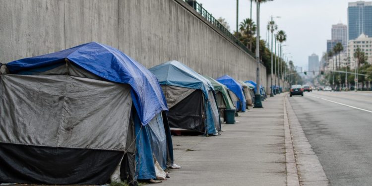 Row of tents along a sidewalk beside a concrete freeway retaining wall in San Diego in neutral daylight, seen with a long lens down the block.