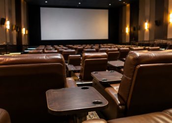 Rows of empty plush reclining seats with small dining tables in a dim cinema auditorium, facing a large blank screen.