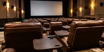 Rows of empty plush reclining seats with small dining tables in a dim cinema auditorium, facing a large blank screen.