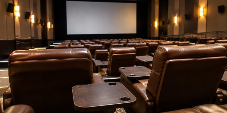 Rows of empty plush reclining seats with small dining tables in a dim cinema auditorium, facing a large blank screen.