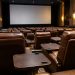 Rows of empty plush reclining seats with small dining tables in a dim cinema auditorium, facing a large blank screen.