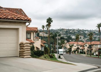 Sandbags stacked beside a garage door on a Southern California residential street under an overcast sky, with hillsides blurred in the background.
