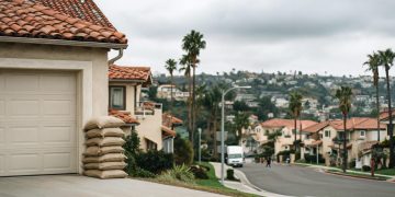 Sandbags stacked beside a garage door on a Southern California residential street under an overcast sky, with hillsides blurred in the background.