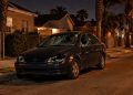 Sedan with a shattered windshield, broken headlights and smashed driver's-side window parked on a narrow South Los Angeles residential street at night.