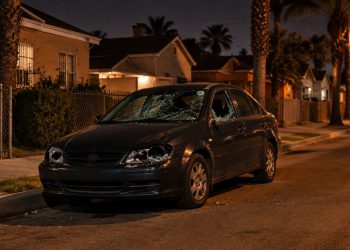 Sedan with a shattered windshield, broken headlights and smashed driver's-side window parked on a narrow South Los Angeles residential street at night.