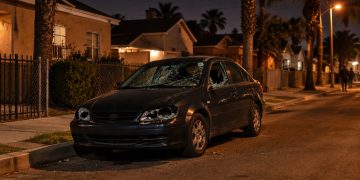 Sedan with a shattered windshield, broken headlights and smashed driver's-side window parked on a narrow South Los Angeles residential street at night.